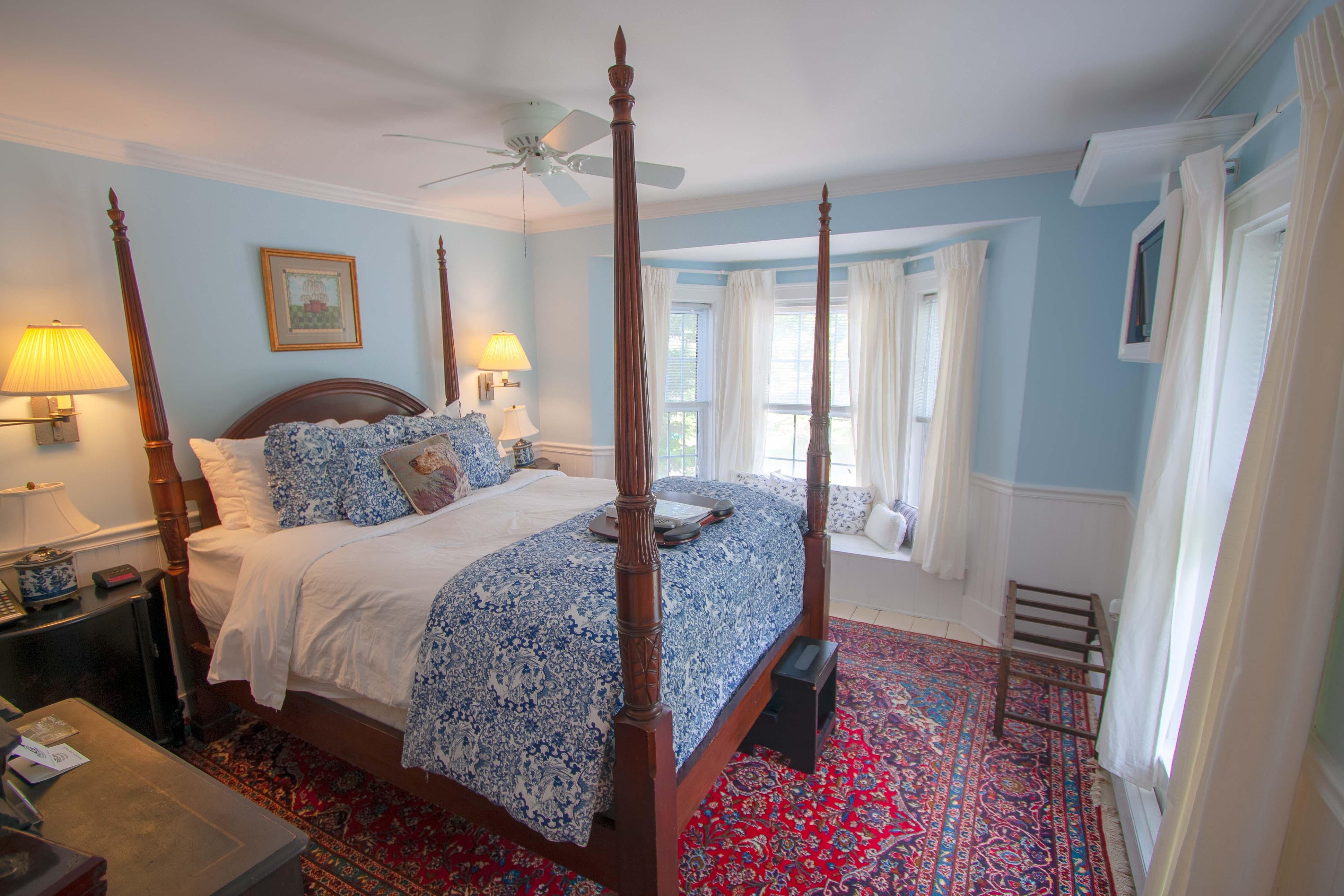 A bright bedroom with light blue walls, a four-poster wood bed with blue floral bedding, a bay window with a window seat, and a red patterned area rug.