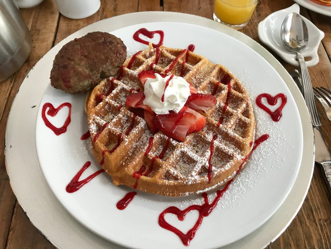 A plate of heart-decorated waffles topped with strawberries, whipped cream, and a sausage patty.