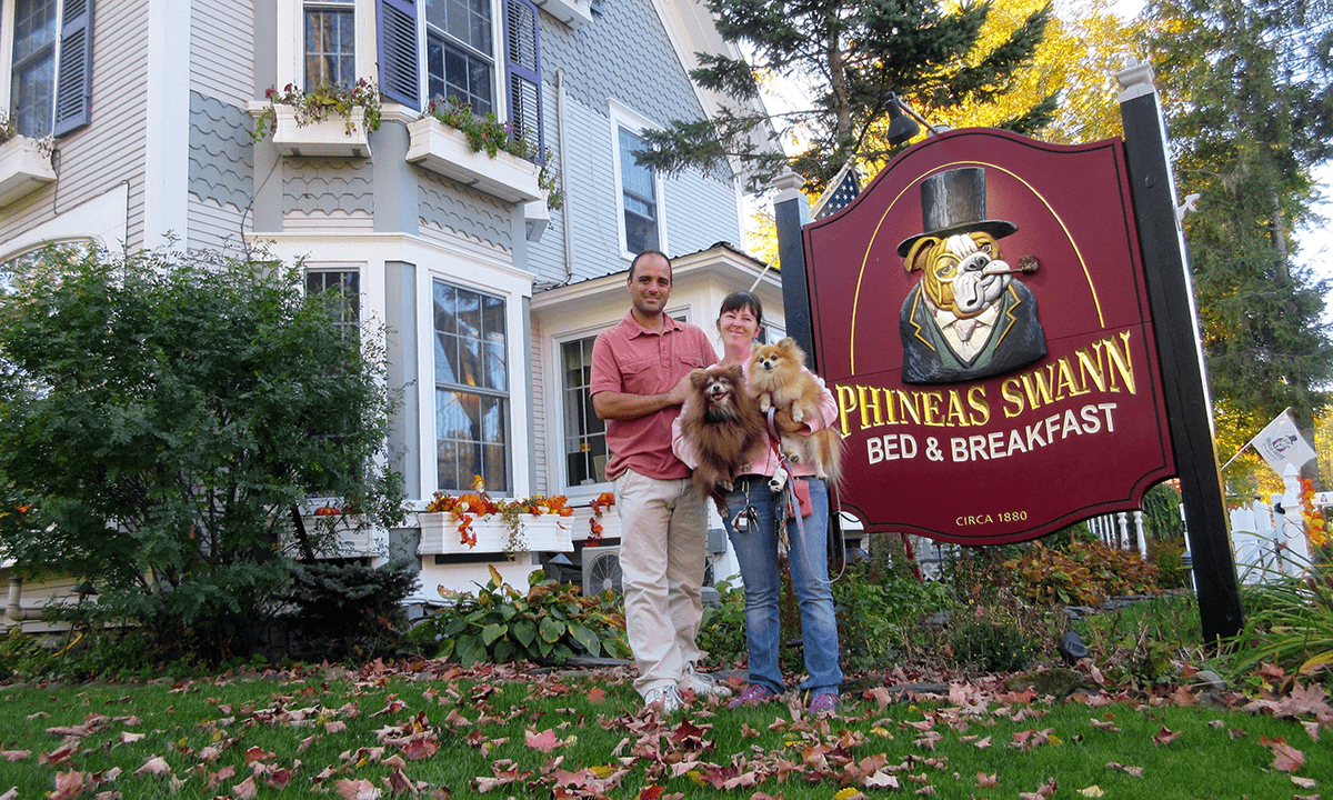 A couple stands with two dogs in front of the Phineas Swann Bed & Breakfast sign.