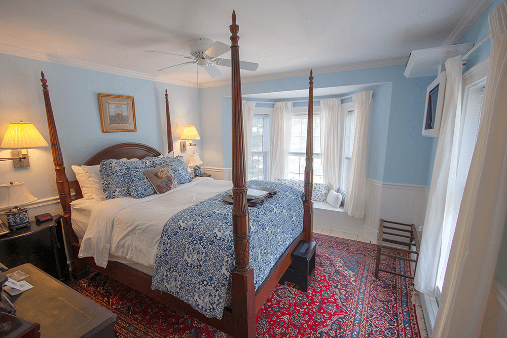 A cozy bedroom featuring a four-poster bed, blue walls, and patterned rugs, with natural light streaming through windows.