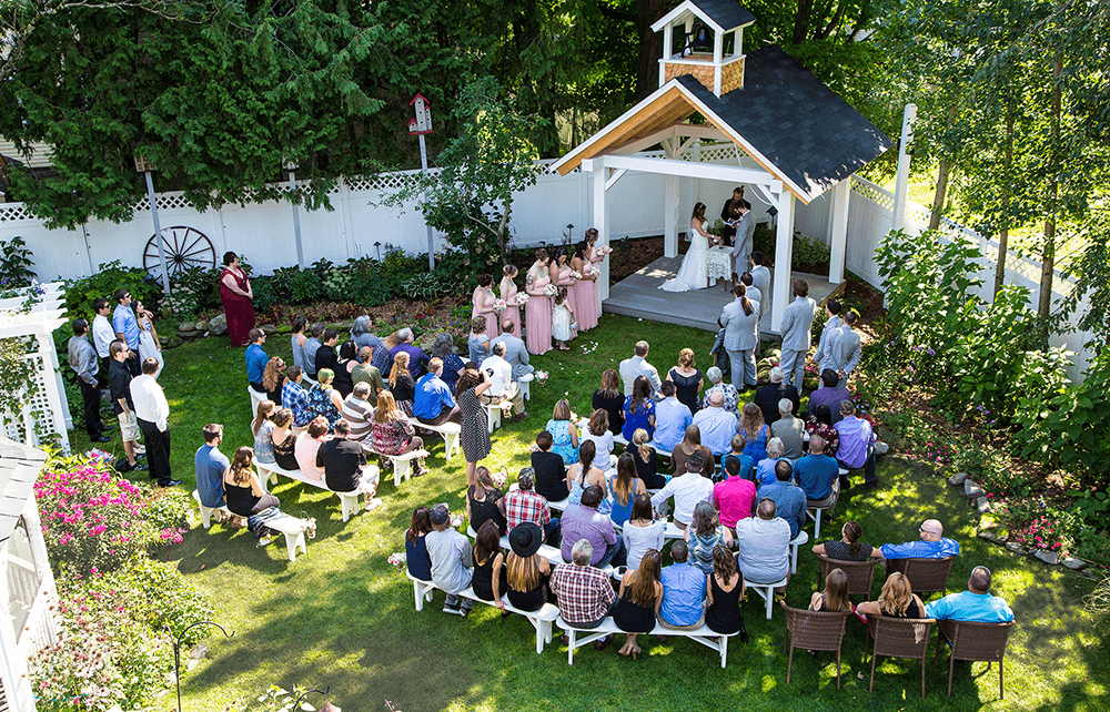 A large gathering of guests watches a couple exchange vows under a gazebo in a garden setting.