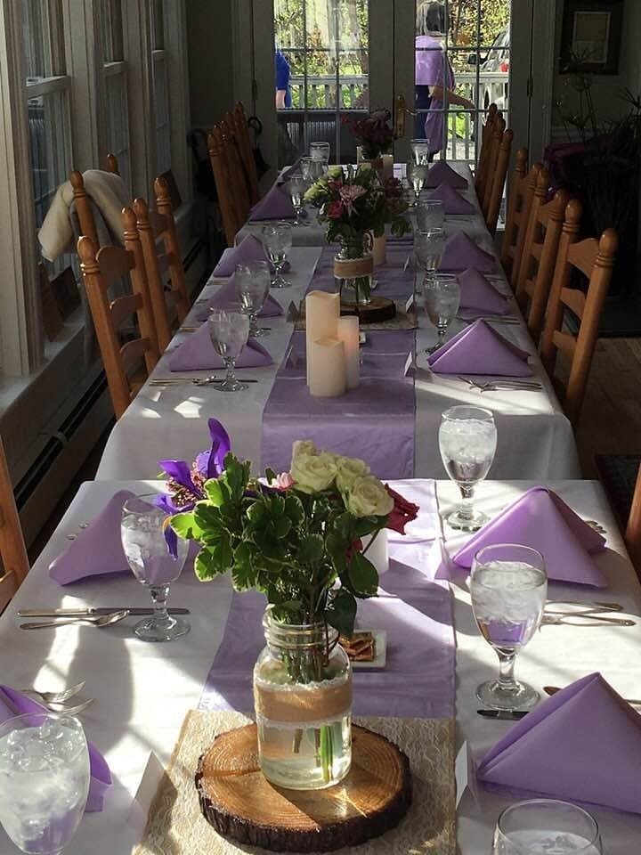 A long dining table is elegantly set with purple napkins, water glasses, floral centerpieces, and candles in a bright room.