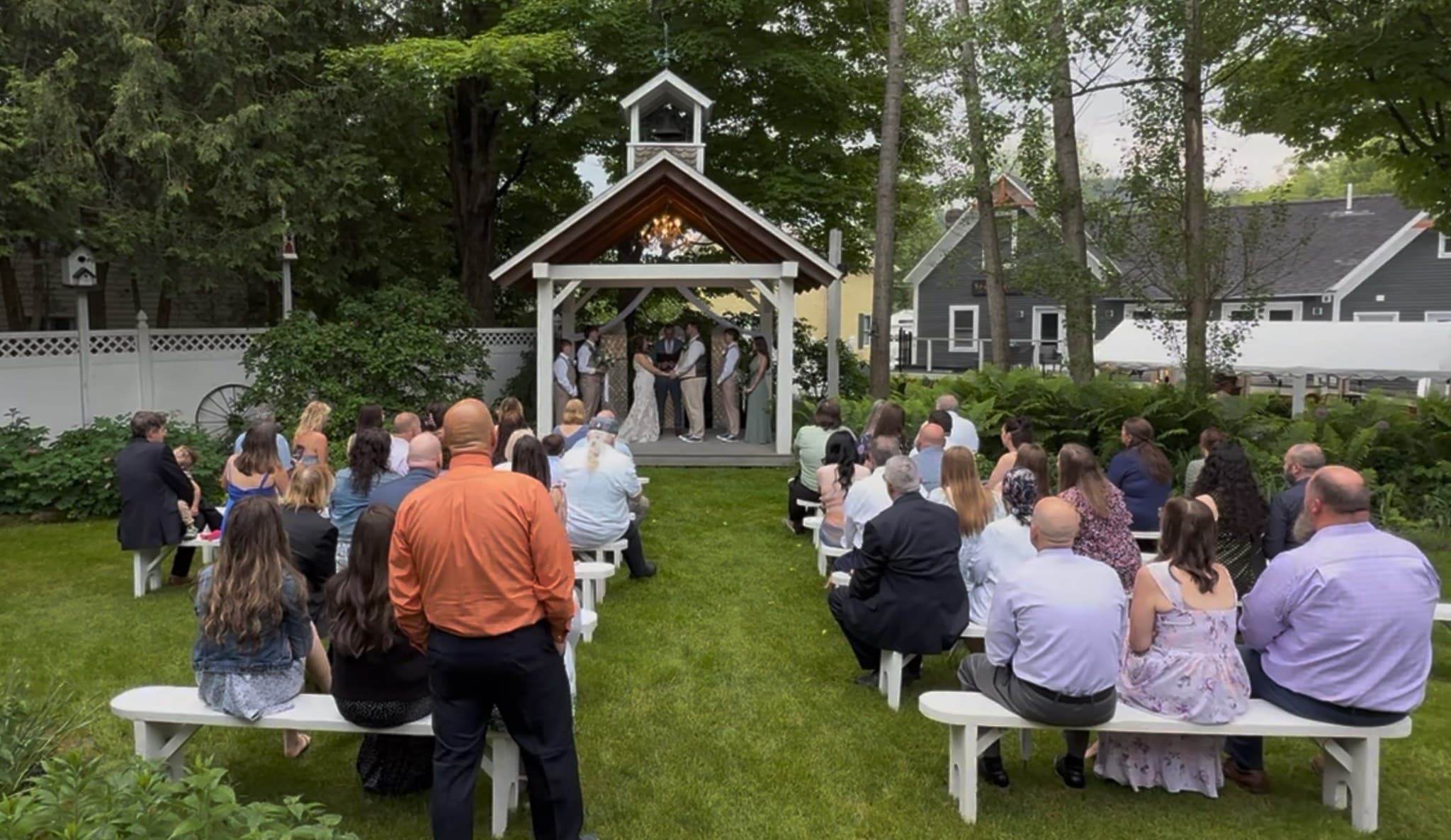 A wedding ceremony taking place outdoors under a gazebo, with guests seated on white benches.