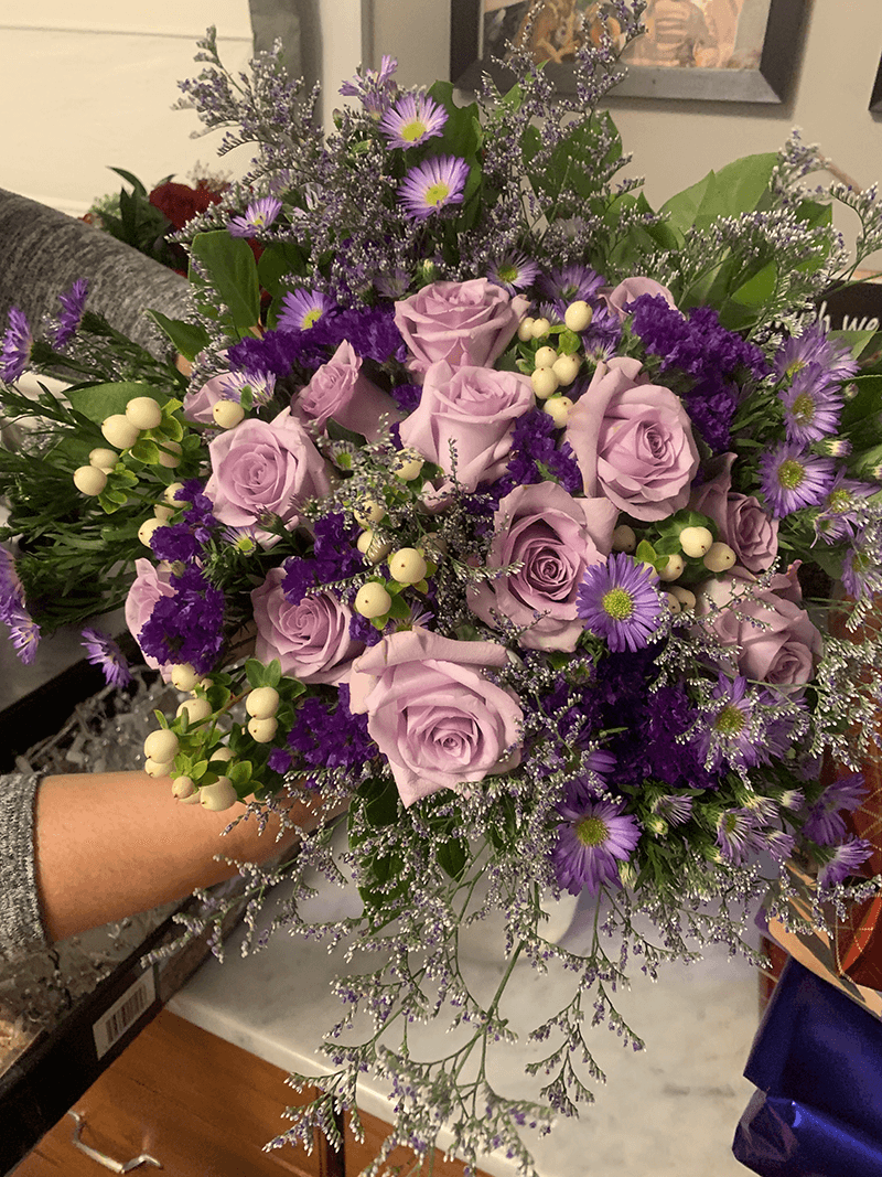 A hand holds a vibrant bouquet featuring lavender roses, purple flowers, and green foliage.