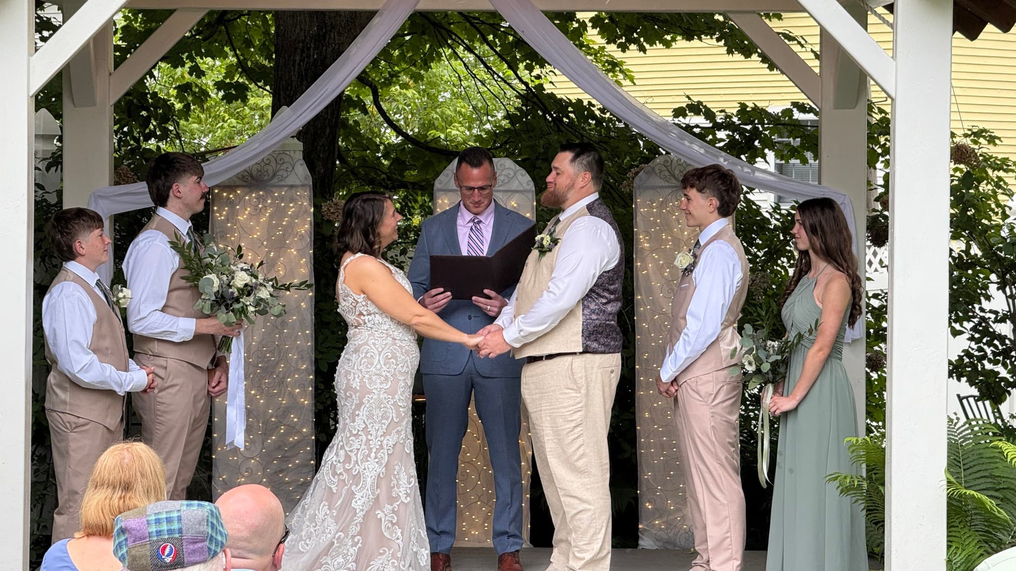 A couple exchanges vows during their wedding ceremony under a decorated gazebo surrounded by their wedding party.