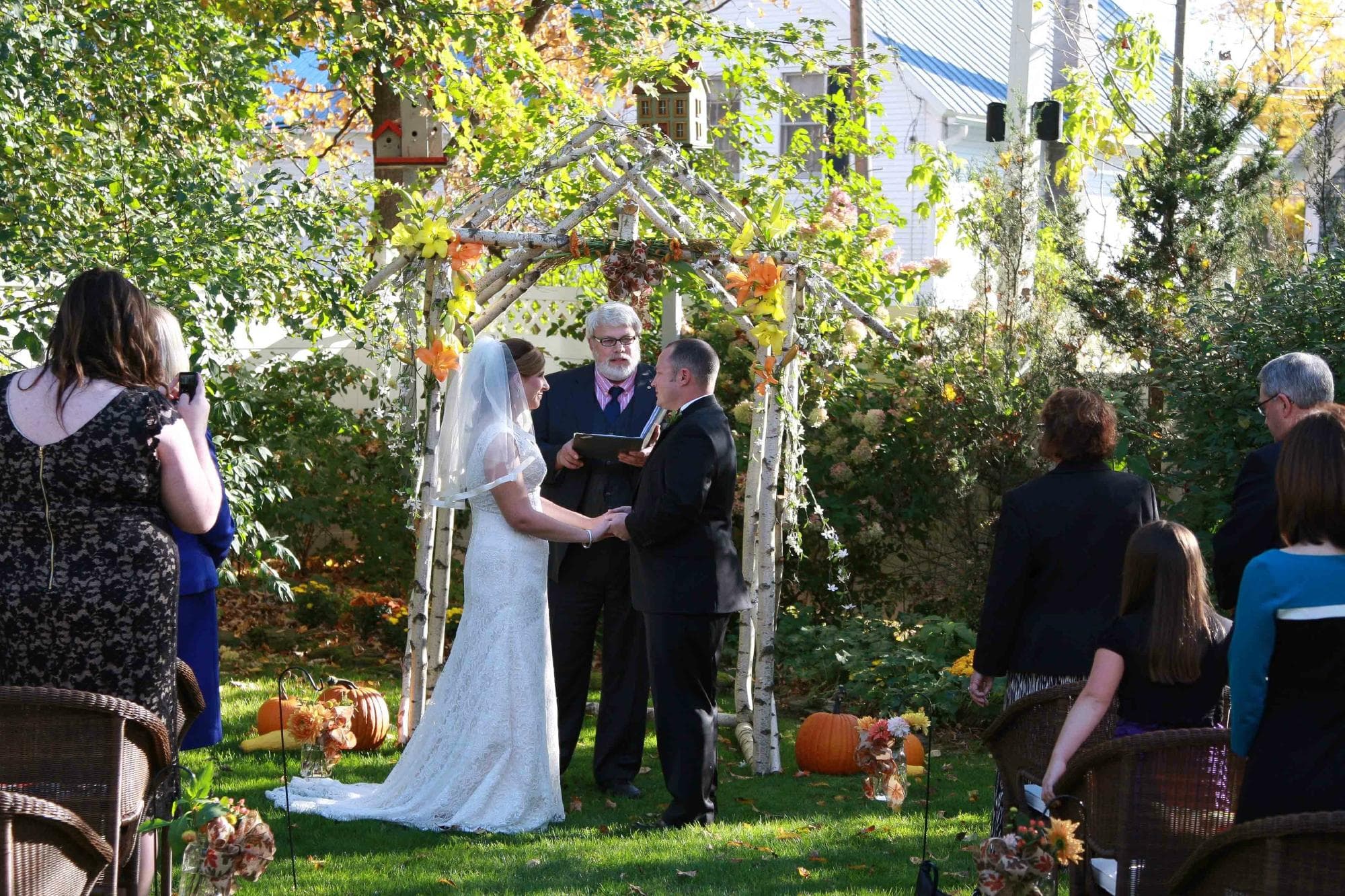 A bride and groom exchange vows under an arch adorned with autumn leaves in a garden ceremony.
