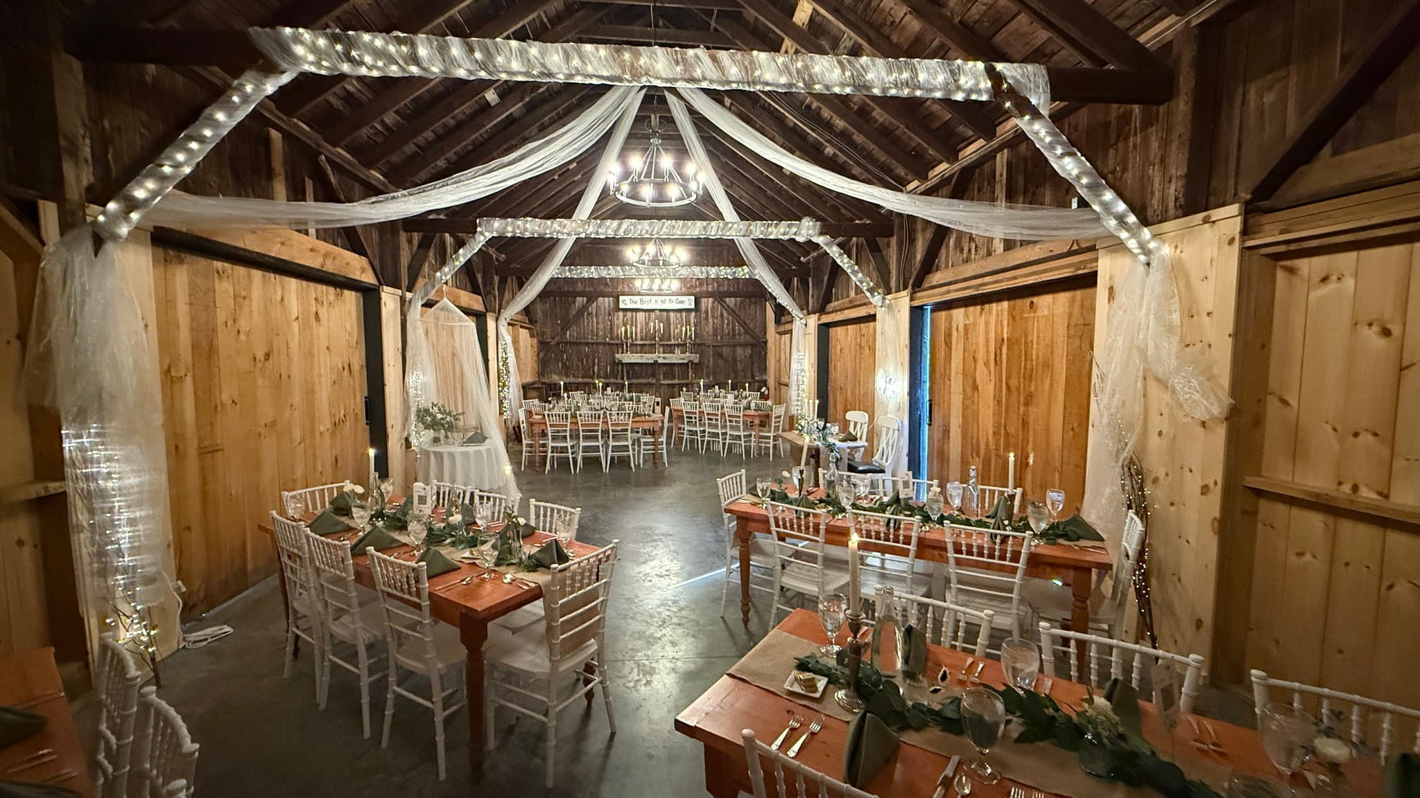 A rustic barn interior is elegantly decorated for a formal event, featuring tables with greenery and soft lighting.